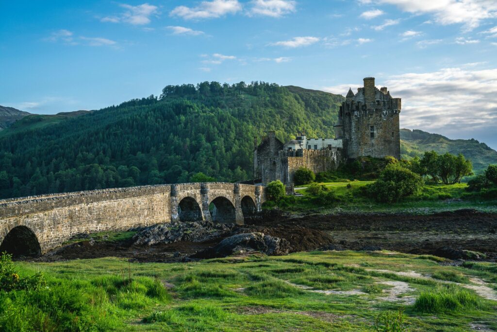 Ancient stone castle with bridge surrounded by lush greenery and rolling hills.